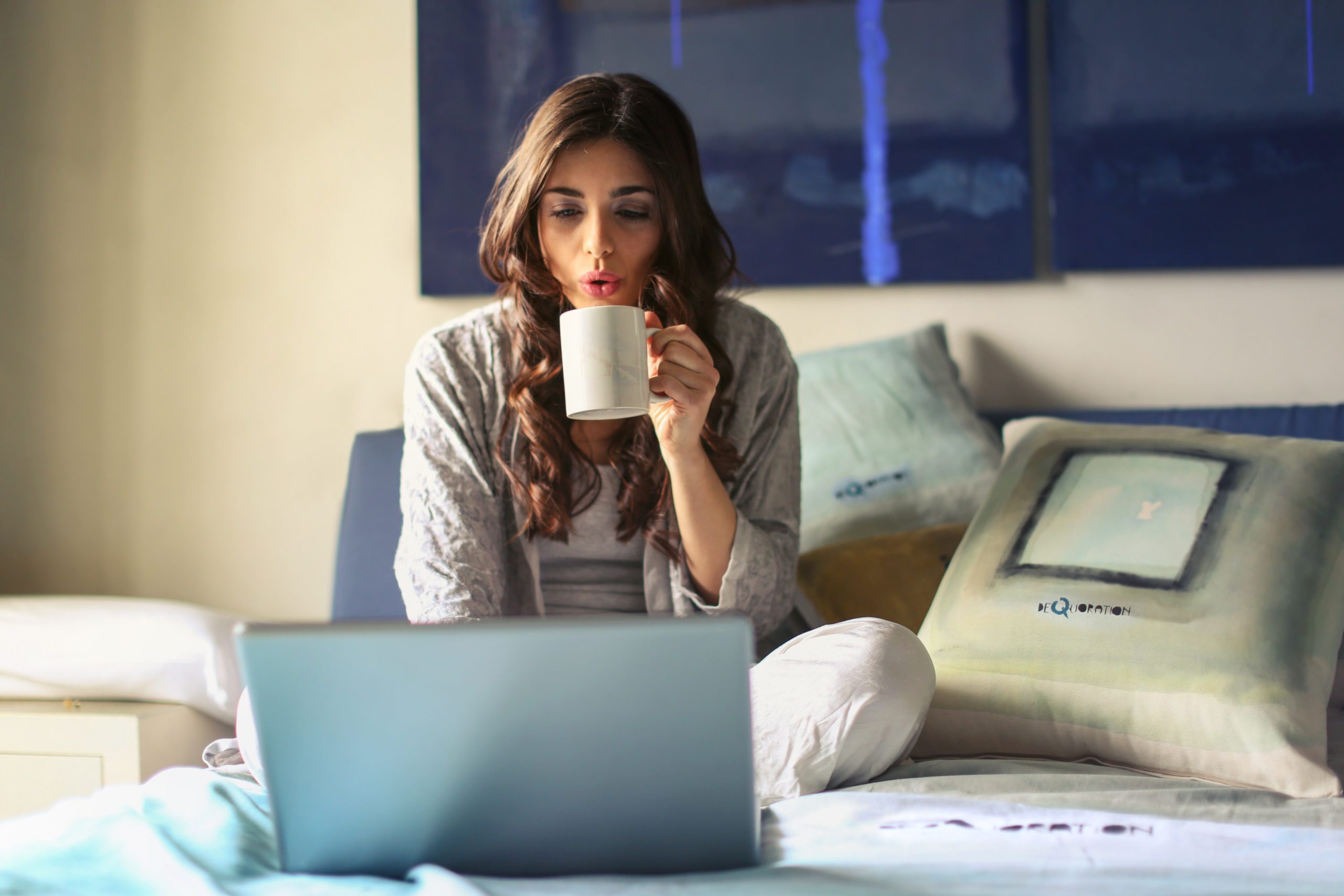 A young woman cools off her cup of coffee while she sits on her bed and logs into her Hoffman Renters Portal, seeking storm advice.