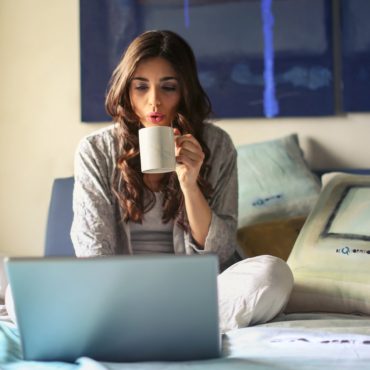A woman working on her laptop on top of her bed, blows on a cup of coffee to cool it off, representative of a homeowner accessing Hoffman Realty's landlord resources