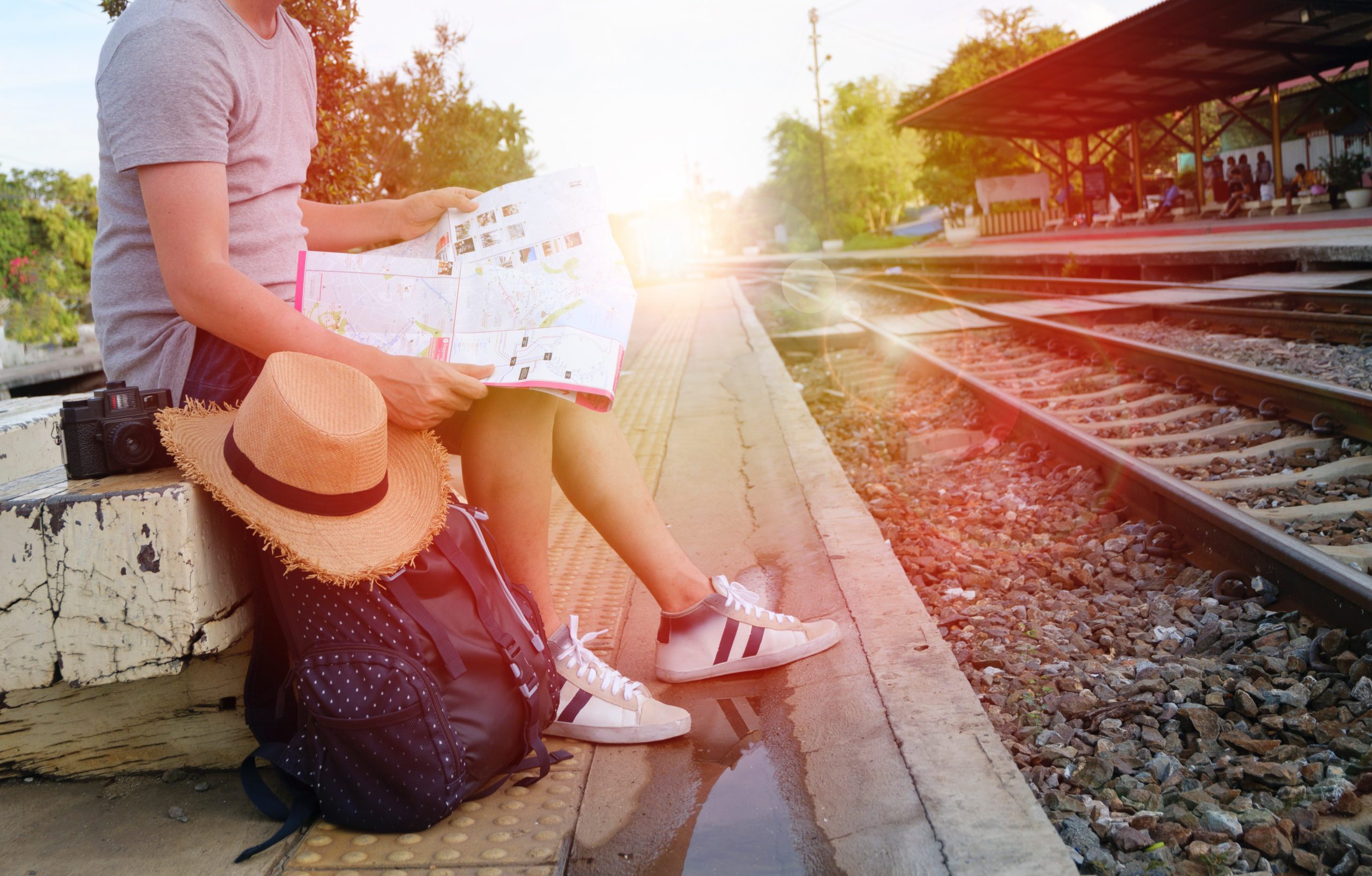 a man sitting near a train rails with his bagpack, camera and hat beside him, while holding a map a man sitting near a train rails with his bagpack, camera and hat beside him, while holding a map