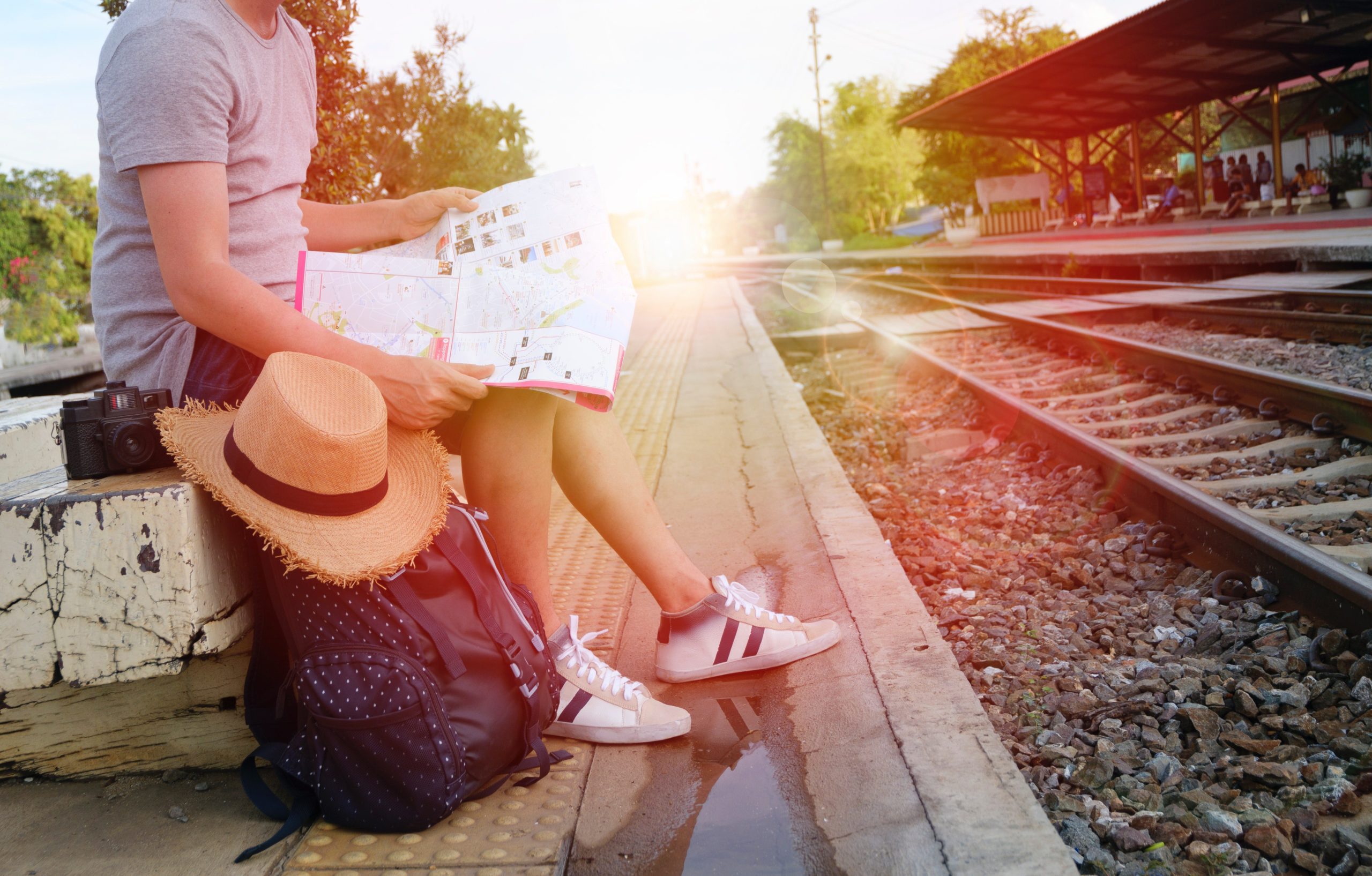 a man sitting near a train rails with his bagpack, camera and hat beside him, while holding a map